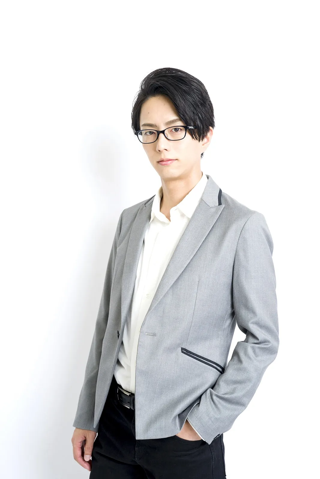A young Asian man with dark hair styled in a short, neat cut, wearing thin-framed glasses, is captured in a waist-up studio shot against a plain white background. He's dressed in a light gray blazer with subtle dark gray detailing along the seams, worn over a crisp white collared shirt and black trousers. His expression is serious and direct, with his gaze meeting the viewer's. One hand is casually in his pocket, conveying a relaxed yet professional demeanor. The overall style of the photograph is clean, simple, and professional, typical of a portrait for a portfolio or publicity purposes.
