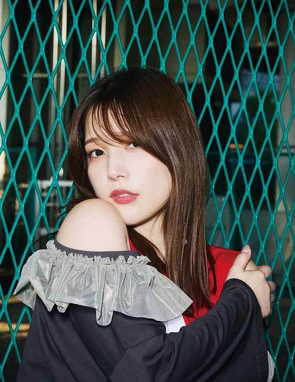 Close-up portrait of a young woman with shoulder-length brown hair, styled with a fringe, posing against a teal diamond-patterned wire mesh fence. She's wearing a black, off-the-shoulder top with a delicate, light grey ruffled trim at the neckline.  Underneath, a hint of red and white is visible.  Her arms are folded across her body, gently holding onto her upper arms, and she looks directly at the camera with a soft, slightly serious expression. Her makeup is natural and understated. The background is blurred but shows the continuation of the wire fence, suggesting an outdoor setting, possibly an urban or industrial area. The image style is contemporary and fashion-focused, with a slightly moody atmosphere.