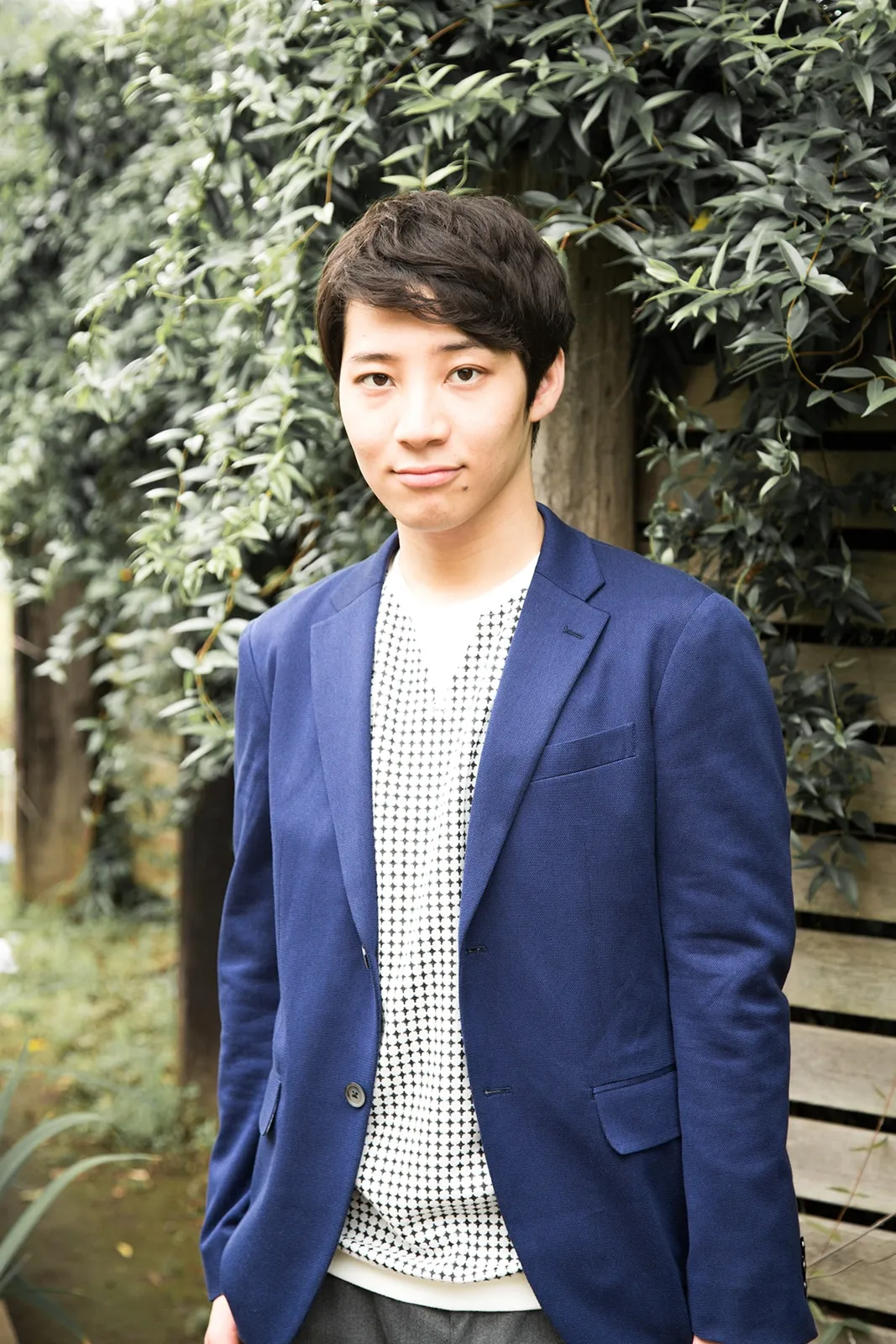 A young Asian man with short, dark brown hair stands slightly angled to the left, looking directly at the camera with a calm expression.  He's wearing a dark blue blazer over a white shirt with a small, repeating black geometric pattern. The shirt is visible at the collar and beneath the blazer's open front. His pants appear to be dark gray. The background is softly blurred, showing a wall partially obscured by lush green foliage with small, oval leaves. The overall style of the image is a clean, simple portrait with a slightly natural feel.