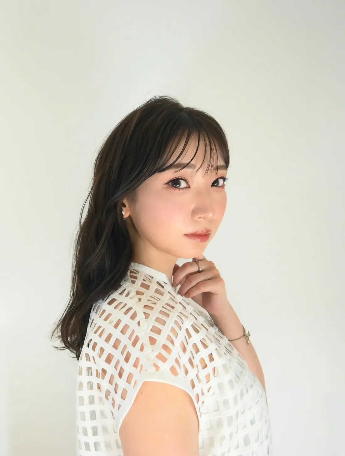 Close-up portrait of a young woman with shoulder-length dark brown hair styled with bangs, glancing over her shoulder. She's wearing a white short-sleeved top with a geometric cut-out pattern, and a delicate silver bracelet on her left wrist.  Her hand rests gently on her chin.  The background is a plain, off-white wall. The image style is a clean, bright portrait photograph, possibly for a fashion or beauty context.